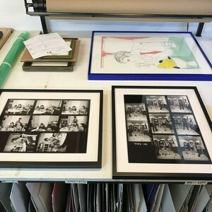 May include: A close-up of a workspace with two framed black and white photographs of a group of people. The photographs are on a white surface with a roll of paper and a framed drawing above them. The photographs are labeled with numbers.