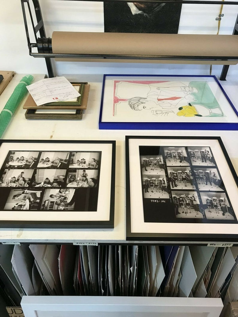 May include: A close-up of a workspace with two framed black and white photographs of a group of people. The photographs are on a white surface with a roll of paper and a framed drawing above them. The photographs are labeled with numbers.