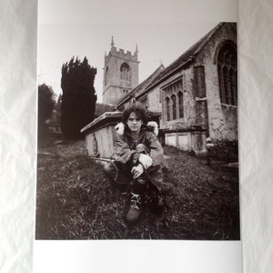 May include: A black and white photograph of a man sitting in front of a church. The man is wearing a jacket and has his arms crossed. The church is in the background and has a tall steeple.