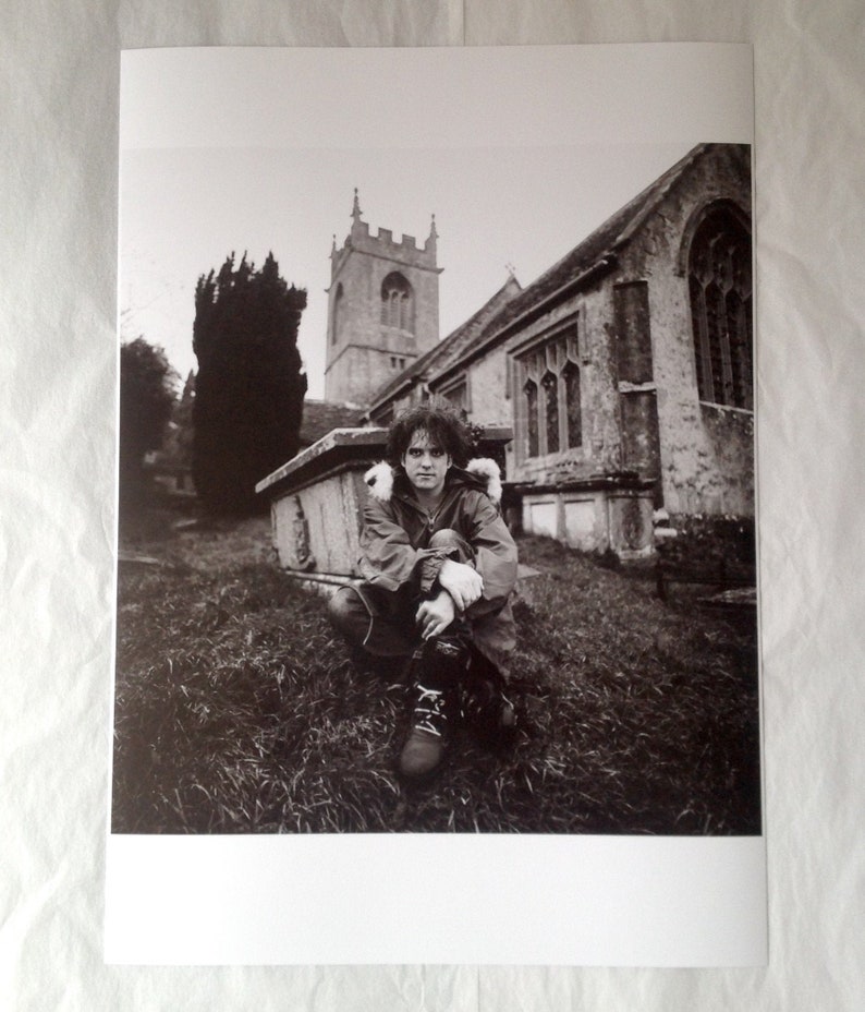 May include: A black and white photograph of a man sitting in front of a church. The man is wearing a jacket and has his arms crossed. The church is in the background and has a tall steeple.