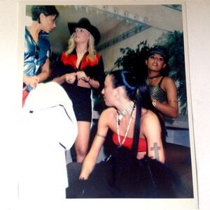 May include: Four women pose for a photo. One woman wears a cowboy hat and a red and black top. Another woman wears a black top with silver accents. The woman in the center wears a red top and a black skirt. The woman on the left wears a blue top and a white shirt.