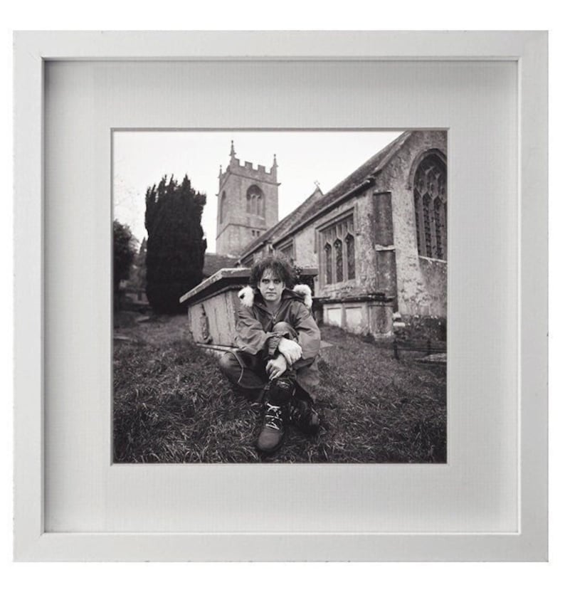 May include: A black and white photograph of a person sitting in front of a church. The person is wearing a jacket and has their arms crossed. The church is in the background and has a tall steeple.