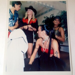 May include: Four women in a room, one wearing a cowboy hat and a red and black shirt, another wearing a black top and a silver necklace, and another wearing a black top with a cross tattoo on her arm.