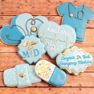 Op de afbeelding: Een set van negen blauwe en gouden gedecoreerde suikerkoekjes met medische thema's. De koekjes bevatten een stethoscoop, een hart met een hartslaglijn, een pil, een verband, een medisch symbool, en de tekst "Doctors Have Heart" en "Congrats Dr. Hart Emergency Medicine".