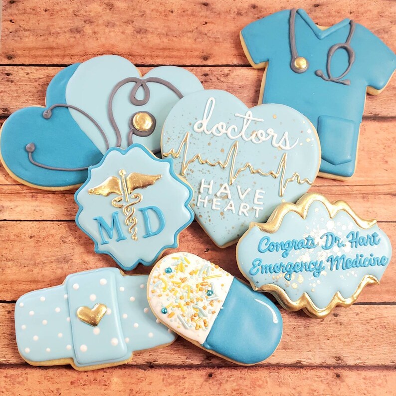 Op de afbeelding: Een set van negen blauwe en gouden gedecoreerde suikerkoekjes met medische thema's. De koekjes bevatten een stethoscoop, een hart met een hartslaglijn, een pil, een verband, een medisch symbool, en de tekst "Doctors Have Heart" en "Congrats Dr. Hart Emergency Medicine".