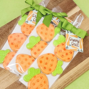 May include: Packaged carrot-shaped cookies with green icing leaves and orange icing dots. Each package is tied with a green ribbon and a tag that reads "Bunny Bait". The cookies are displayed on a wooden cutting board.