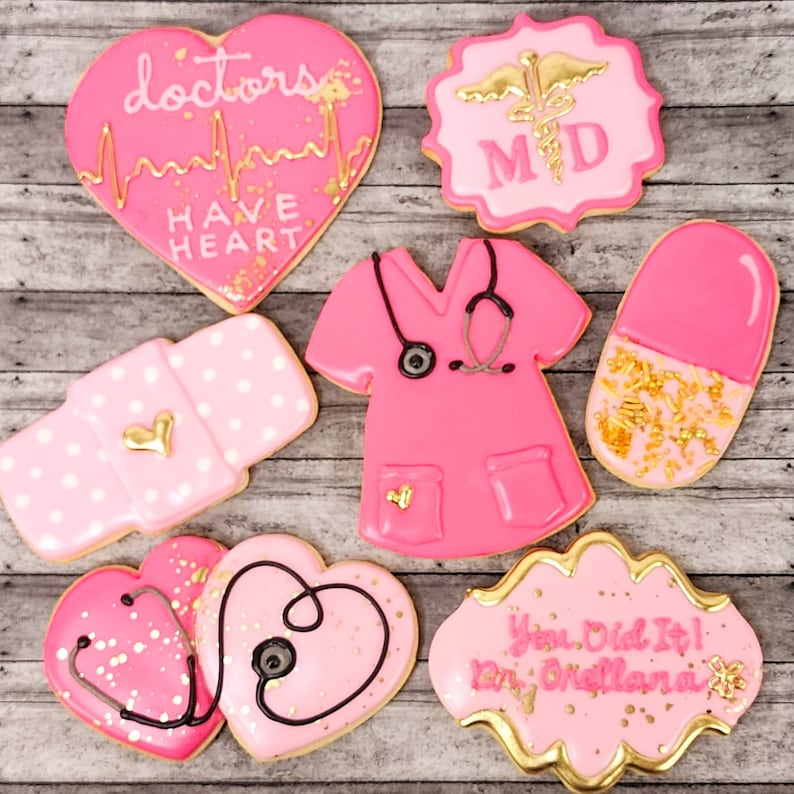 Op de afbeelding: Roze en gouden gedecoreerde suikerkoekjes met medische thema's. De koekjes bevatten een hart met de tekst "doctors have heart", een stethoscoop, een pil, een verband, een scrubtop en een bord met de tekst "You Did It! Dr. [Name]".