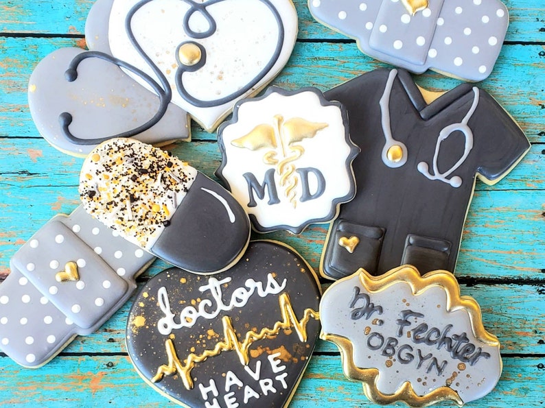 Op de afbeelding: Assortiment van gedecoreerde suikerkoekjes met een medisch thema. De koekjes tonen een stethoscoop, een pil, een verband, een hart met een hartslag, een doktersjas en een bord met de tekst "MD".