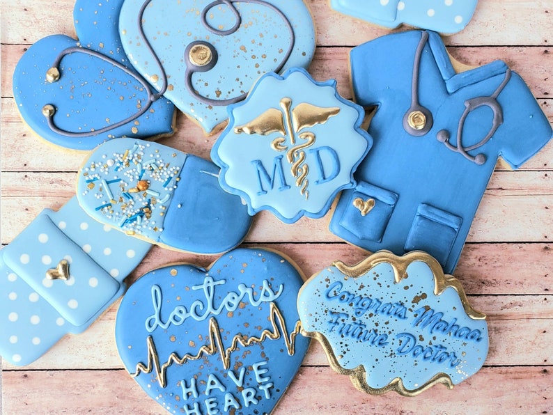 Op de afbeelding: Assortiment van blauwe en gouden gedecoreerde suikerkoekjes met verschillende medische thema's. De koekjes bevatten een stethoscoop, een pil, een hart met de tekst "doctors have heart", een medisch scrubtop en een koekje met de tekst "Congrats Mahaa Future Doctor".