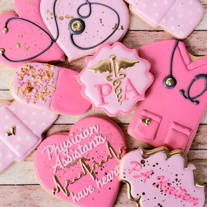 Op de afbeelding: Roze en gouden gedecoreerde suikerkoekjes met verschillende ontwerpen, waaronder een hart met een stethoscoop, een pil, een scrubtop en een hart met de tekst "Physician Assistants have heart".
