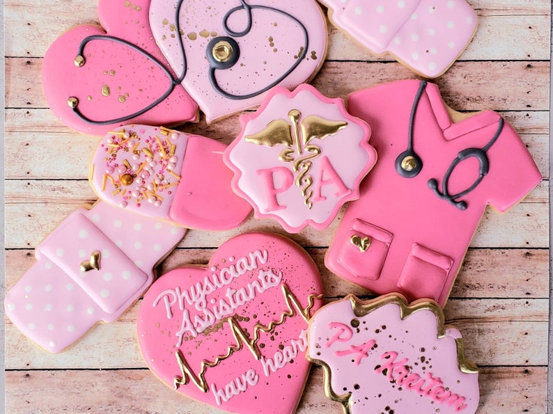 Op de afbeelding: Roze en gouden gedecoreerde suikerkoekjes met verschillende ontwerpen, waaronder een hart met een stethoscoop, een pil, een scrubtop en een hart met de tekst "Physician Assistants have heart".