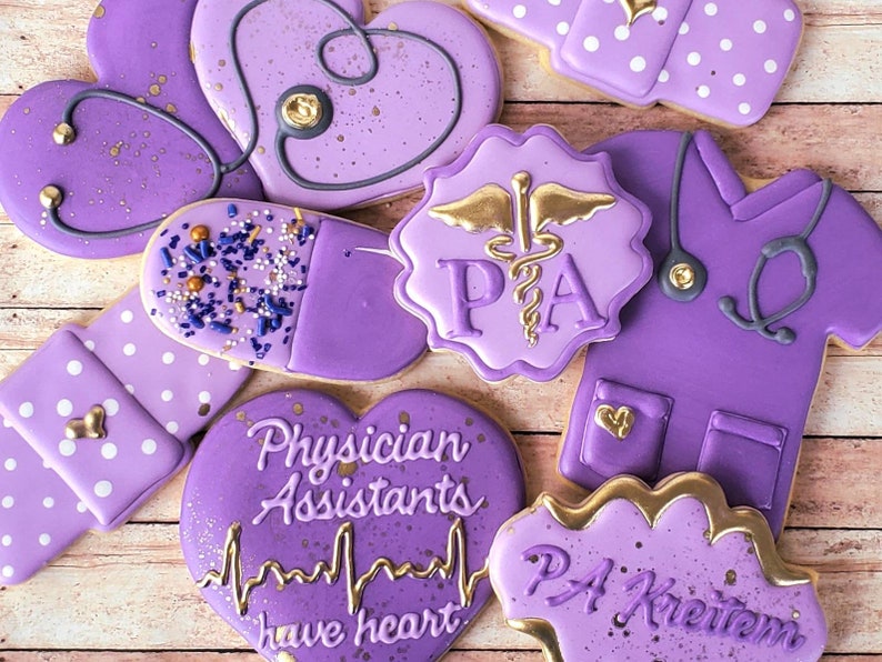 Op de afbeelding: Paarse en gouden gedecoreerde suikerkoekjes met verschillende ontwerpen die betrekking hebben op het medische beroep. De koekjes bevatten een stethoscoop, een pil, een hart en de letters "PA" voor Physician Assistant. Een koekje heeft de tekst "Physician Assistants have heart" en een hartslaggrafiek.