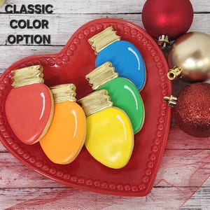 May include: Five Christmas light-shaped cookies decorated with red, orange, yellow, green, and blue icing. The cookies are arranged on a red heart-shaped plate. The text "CLASSIC COLOR OPTION" is visible in the top left corner of the image.