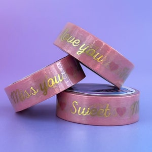 May include: Three rolls of pink washi tape with gold lettering. The top roll says "Love you so", the middle roll says "Miss you", and the bottom roll says "Sweetest Miss".