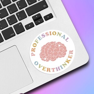 May include: A round sticker with the words "PROFESSIONAL OVERTHINKER" in pastel colors encircling a pink brain graphic. The sticker is placed on a silver laptop keyboard against a gradient background of purple and blue.