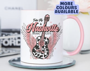 May include: White ceramic mug with a pink rim and handle. The mug features a pink and black cow print guitar with wings and the text "Take Me to Nashville Music City".