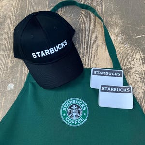 May include: A green apron with the Starbucks logo and the words "Starbucks Coffee" printed on it. There is a black baseball cap with the word "Starbucks" printed on it and two white name tags with the word "Starbucks" printed on them.