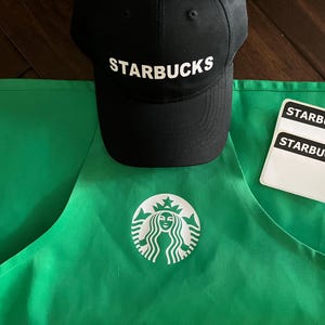 May include: A green apron with the Starbucks logo, a black cap with "STARBUCKS" in white, and two name tags. The apron has a large white logo on the front. The cap is black with a curved brim.