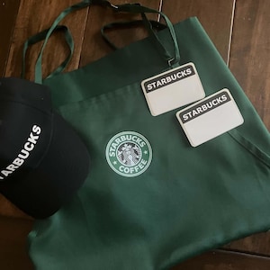 May include: A black baseball cap with the word STARBUCKS embroidered in white, paired with a dark green apron featuring the circular Starbucks Coffee logo in the center and two white rectangular name tags with the word STARBUCKS printed on them.