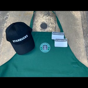 May include: A black baseball cap and a green apron with the Starbucks logo. The cap has "Starbucks" in white lettering. The apron has the Starbucks logo and two name tags with the Starbucks logo.