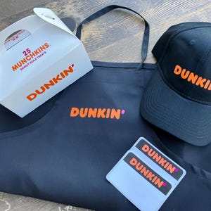 May include: A black apron, baseball cap, and stickers with the Dunkin' logo in orange. A white box with the text "25 Munchkins Donut Hole Treats" and the Dunkin' logo is also present. The items are arranged on a wooden surface.