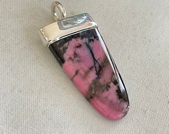 May include: A silver pendant featuring a pink and black rhodochrosite stone. The stone is cut in a long, pointed shape and has a silver bail at the top.