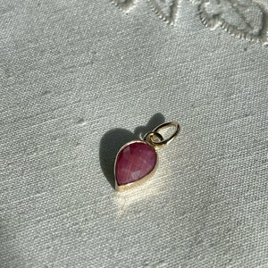 以下が含まれることがあります： しずく型のピンク色の宝石が付いたゴールドのペンダント。ペンダントの上部にはチェーン用の小さなループが付いています。宝石は金のベゼルにセットされています。ペンダントは白く、質感のある表面に表示されています。