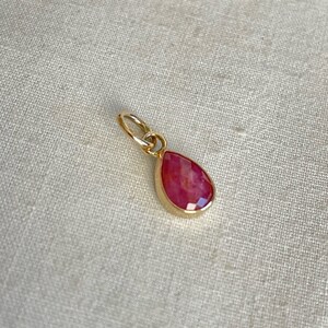 May include: A teardrop-shaped pendant with a faceted, reddish-pink gemstone set in a gold-colored bezel. The pendant has a small, gold-colored loop at the top for attaching to a chain. The background is a light beige fabric.