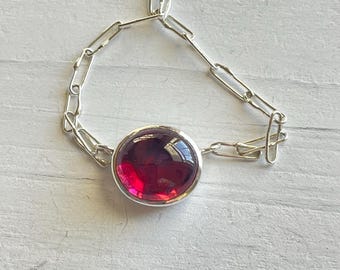 May include: A silver chain bracelet with a large, round, red gemstone.