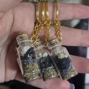 May include: Three small glass bottles filled with herbs and crystals, each with a cork stopper and attached to a gold keychain. The bottles contain layers of green, purple, black, and white materials.