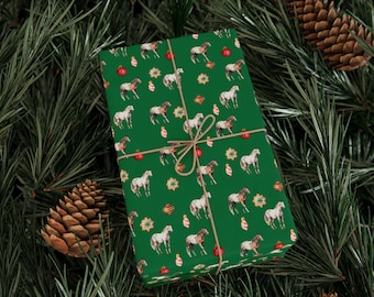 Puede incluir: Regalo envuelto con un fondo verde con un patrón de caballos blancos y marrones, adornos rojos y galletas doradas. El regalo está atado con hilo y colocado sobre un fondo de agujas de pino y piñas.