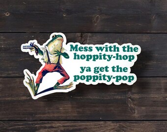 May include: A die-cut sticker featuring a green frog wearing a white shirt and red pants with suspenders, holding a small black pistol. The text reads, "Mess with the hoppity-hop ya get the poppity-pop" in dark green lettering on a white background.