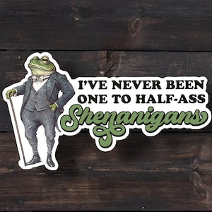 May include: A sticker featuring a cartoon frog wearing a suit and holding a cane. The text on the sticker reads "I've never been one to half-ass shenanigans."