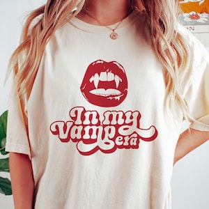 Könnte beinhalten: Ein weißes T-Shirt mit einem roten Aufdruck eines Vampirmundes mit Zähnen und dem Text "In my Vampire Era".