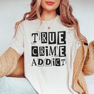 May include: A white t-shirt with the text "TRUE CRIME ADDICT" printed in black letters, with each letter in a separate box.