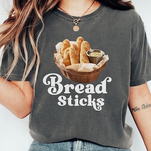 May include: A charcoal grey t-shirt with a white graphic of breadsticks in a basket with dipping sauce. The text "Bread Sticks" is written in a retro font below the image.