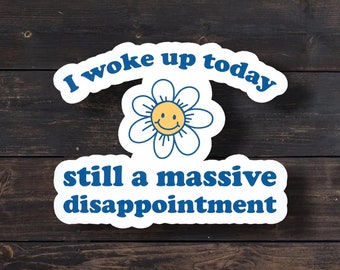 May include: A white die-cut sticker featuring a yellow smiley face flower with blue outlines. The text "I woke up today" is arched above the flower in blue, and the text "still a massive disappointment" is printed below the flower in blue.
