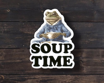 May include: A die-cut sticker featuring a green frog wearing a blue collared shirt, sitting and holding a white bowl of soup. Below the frog, the text SOUP TIME is printed in large, bold, black capital letters against a white background border.