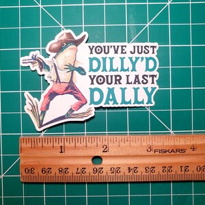May include: A sticker featuring a cartoon frog wearing a cowboy hat and holding a gun. The text on the sticker reads "You've Just Dilly'd Your Last Dally."