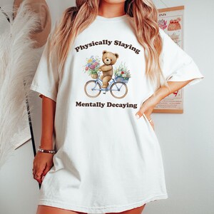 May include: White oversized t-shirt with a brown bear riding a blue bicycle with flowers. The text on the shirt reads "Physically Slaving Mentally Decaying".