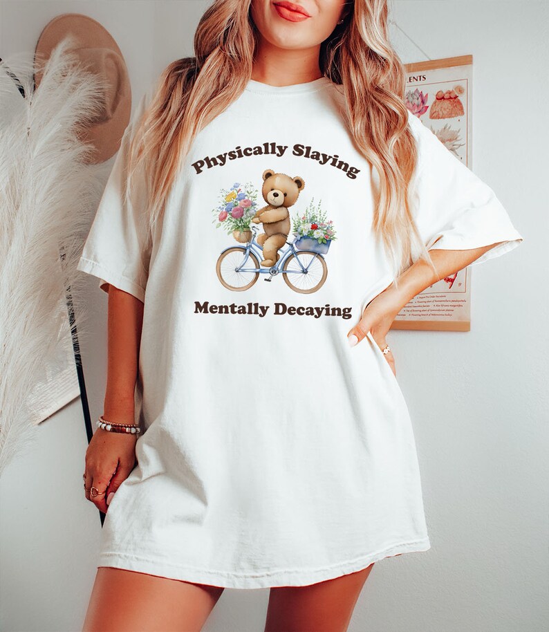 May include: White oversized t-shirt with a brown bear riding a blue bicycle with flowers. The text on the shirt reads "Physically Slaving Mentally Decaying".