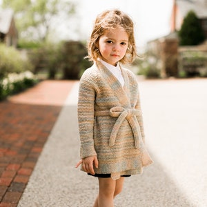 Op de afbeelding: Een jong meisje draagt een gestreepte cardigan met een strikband, een wit overhemd met kraag en bruine schoenen. Ze staat op een pad van bakstenen.