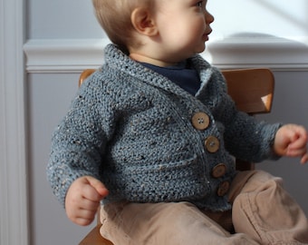 May include: A baby wearing a blue-grey crocheted cardigan with large wooden buttons. The cardigan has a textured appearance and a shawl collar. The baby is also wearing a navy blue shirt and tan pants, sitting on a wooden chair.