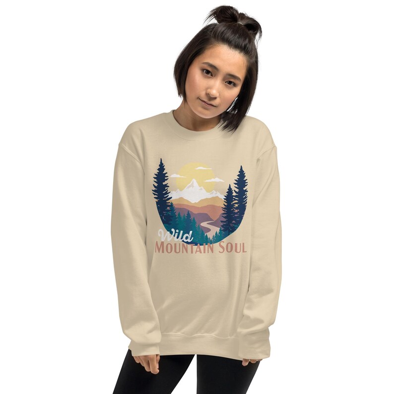 May include: A beige sweatshirt with a graphic of a mountain landscape with a sun and trees. The text "Wild Mountain Soul" is printed below the graphic.