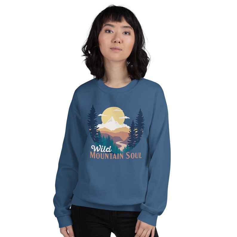 May include: A blue sweatshirt with a graphic of a mountain range, a sun, and trees. The text "Wild Mountain Soul" is printed below the graphic.