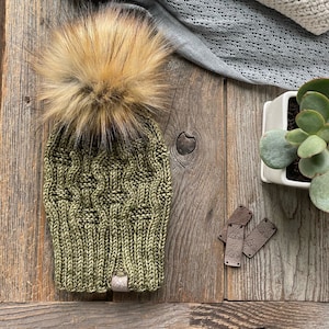 May include: An olive green knit hat with a brown faux fur pom-pom. The hat has a cable knit design and a small tag. Several folded blankets and a succulent plant are in the background.