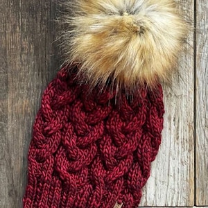May include: A knitted burgundy beanie with a cable knit design and a large, fluffy, faux fur pom-pom in shades of brown and tan. The hat has a small, rectangular tag attached to the brim. The hat is displayed on a wooden surface.