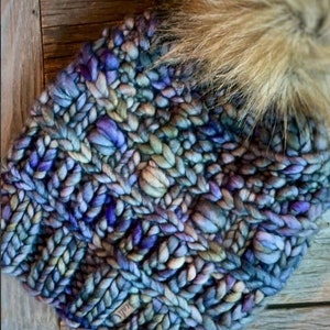 May include: Hand-knitted beanie in shades of blue, purple, and green with a faux fur pom-pom. The chunky knit hat features a textured cable stitch pattern. A small tag with the word "icali" is visible on the side.