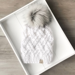 Op de afbeelding: Een witte gebreide muts met een textuurpatroon en een grote, pluizige grijze en witte pompon. De muts heeft een klein label met de tekst "cali". De hoed wordt in een witte bak gepresenteerd.