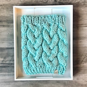 May include: A turquoise cable-knit cowl neck warmer. The chunky knit has a textured, braided design. The cowl is displayed in a white tray on a wooden surface. A small tag with the word "cali" is attached to the cowl.